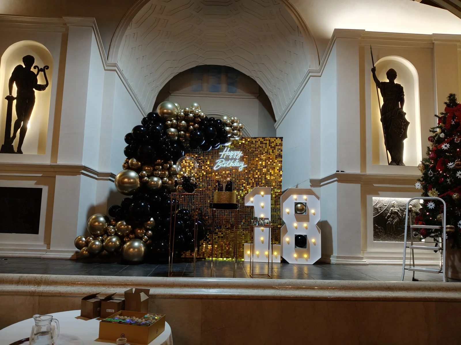 Vista della sala con fondale shimmer dorato, palloncini neri e oro e numero 18 luminoso.