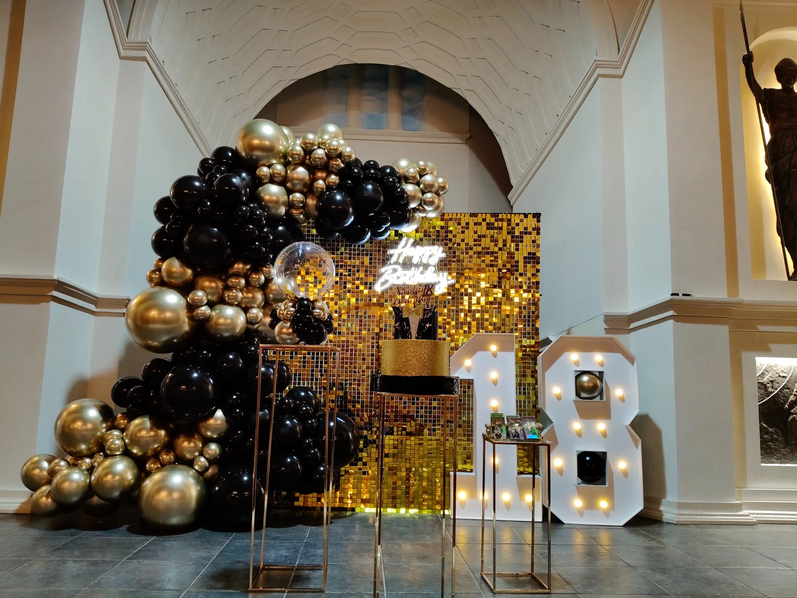 Allestimento per diciottesimo con palloncini neri e oro, fondale dorato e numero 18 luminoso.