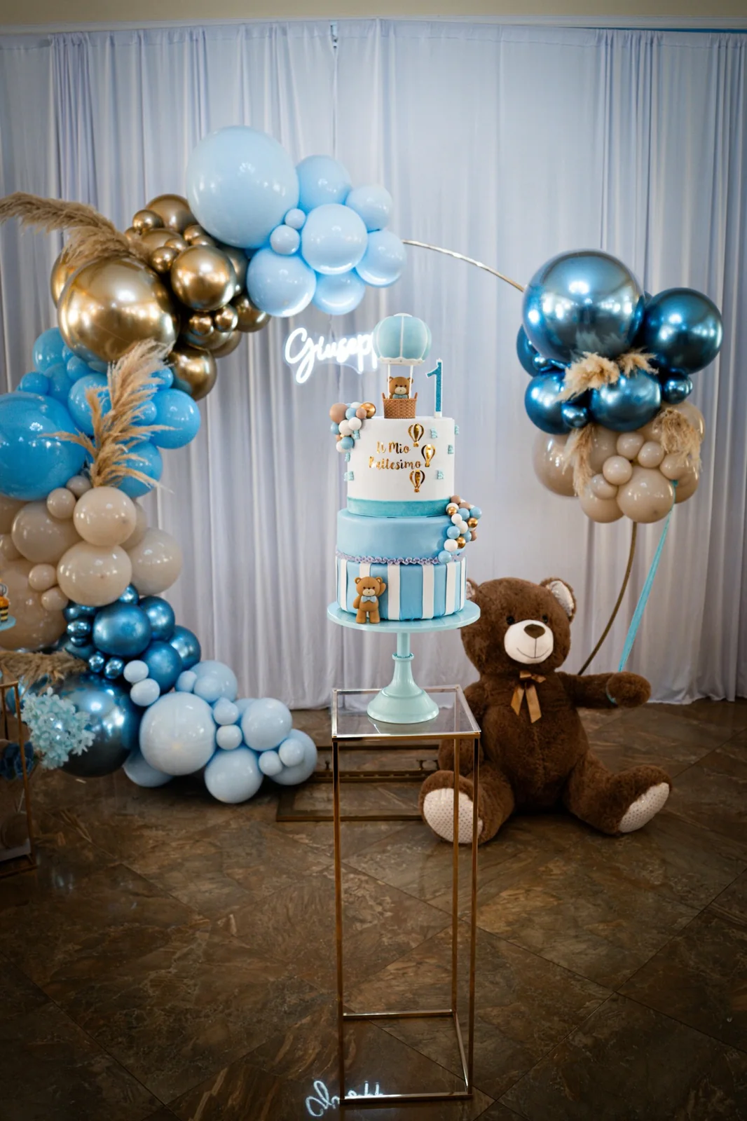 Zona torta per primo compleanno con palloncini azzurri, avorio e oro e un orsetto.