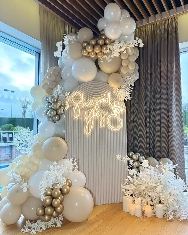 Fondale per proposta di matrimonio con palloncini bianchi e oro, fiori e scritta luminosa.